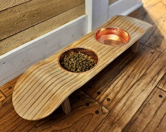 May include: A wooden pet feeder with two copper-lined bowls. One bowl contains dry pet food. The feeder is made of light-colored wood with a curved design and is elevated on small legs. The bowls are circular and have a hammered copper finish.