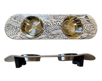May include: A white and black patterned pet bowl holder with two stainless steel bowls. The holder is shaped like a skateboard. The bowls are held in place by a wooden base with black supports.
