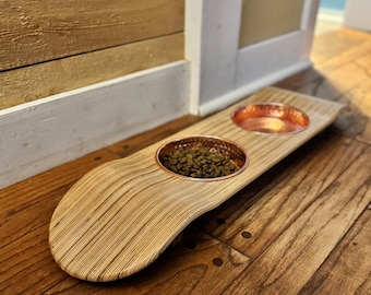May include: A wooden pet bowl with two copper-colored bowls. One bowl is filled with pet food. The wooden base has a curved design and a natural wood grain finish. The bowls are set into the wood.