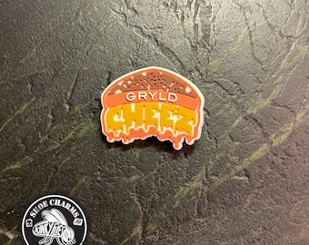 May include: A yellow, orange, and brown shoe charm with the text "GRYLD CHEEZ" in a dripping font. The charm is shaped like a hamburger and is a shoe accessory.