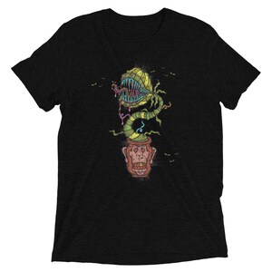Puede incluir: Camiseta negra con una ilustración colorida de una planta carnívora con dientes afilados y un tallo sinuoso. La planta emerge de una maceta con una cara de mono. Pequeños pájaros negros están dispersos alrededor del diseño.