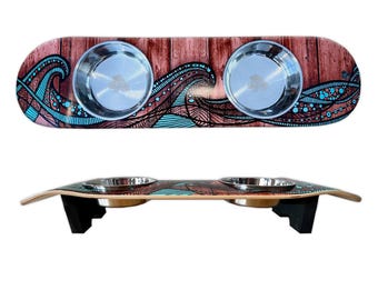 May include: A wooden pet food bowl stand with a blue and brown wave design. The stand has two stainless steel bowls and is elevated on two black legs.