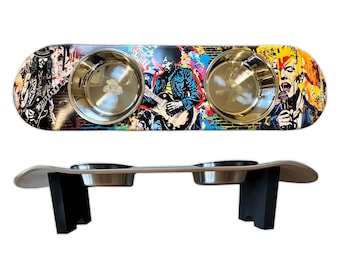 May include: A dog bowl stand made from a repurposed skateboard. The skateboard has a colorful, graffiti-style design featuring rock and roll musicians. Two stainless steel bowls are set into the skateboard, which is supported by two black wooden legs.