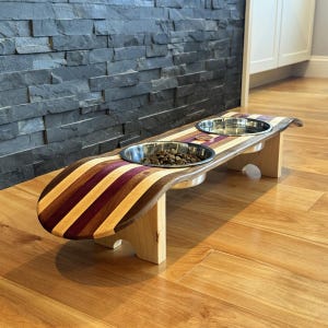 May include: A wooden pet feeder with two stainless steel bowls. The feeder has a curved design with alternating stripes of purple, brown, and tan wood. The feeder is elevated on light-colored wooden legs.