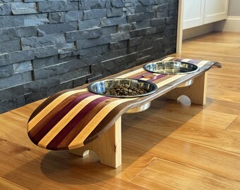 May include: A wooden pet feeder with two stainless steel bowls. The feeder has a curved design with alternating stripes of purple, brown, and tan wood. The feeder is elevated on light-colored wooden legs.