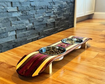 May include: A wooden pet bowl stand with two stainless steel bowls. The stand is made from a repurposed skateboard with a purple, yellow, and brown striped design. One bowl is filled with pet food. The stand is supported by a wooden base.