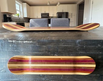 May include: Two wooden skateboards with a striped design. The top skateboard is angled, showing the curved shape. The bottom skateboard is flat, displaying the full striped pattern of various wood tones, including red, purple, and brown.