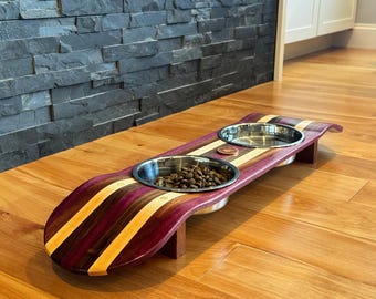 May include: A wooden pet bowl stand with two stainless steel bowls. The stand is made of purple, brown, and yellow wood. One bowl is filled with pet food. The stand is on a hardwood floor.