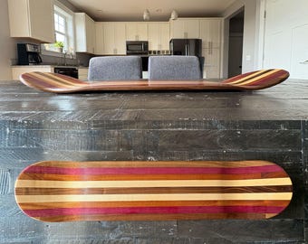 May include: Two wooden skateboards with a striped design. The top skateboard is positioned at an angle, showcasing a curved shape. The bottom skateboard is flat, displaying a colorful pattern of red, brown, and tan stripes. The skateboards are made of wood.