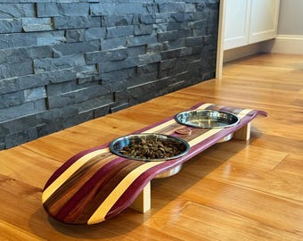 May include: A wooden pet feeder with two stainless steel bowls. The feeder is made from a skateboard deck with alternating stripes of purple, brown, and light wood. The bowls are filled with pet food.