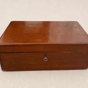 Puede incluir: Una caja rectangular de madera con un acabado liso y pulido en color marrón medio. La caja cuenta con una tapa con bisagras y una pequeña cerradura de metal plateado centrada en el panel frontal, diseñada para cerrar el contenedor de almacenaje.