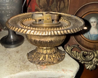 May include: A gold-toned metal table lighter featuring a wide, circular tray base with a beaded rim and a pedestal stand decorated with leaf-like carvings. The lighter mechanism is integrated into the center of the tray, showcasing a metallic finish.