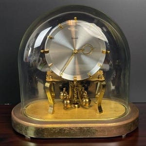 May include: A vintage clock with a silver face and gold-tone accents, encased in a clear glass dome. The clock features a pendulum and is set on a wooden base with a gold-colored trim. The clock's face has the word "Hermle".