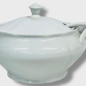 May include: A white ceramic soup tureen with a lid and handles. The tureen has a rounded body and a lid with a small knob. A matching spoon rests on the side. This classic serving dish is perfect for soups and stews.