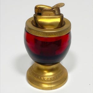 May include: A vintage table lighter with a gold-coloured metal base and top. The body is a deep red glass. The lighter mechanism is gold-coloured. The base has an ornate, detailed design. A collectible item.