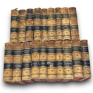 May include: A collection of antique books with tan leather covers and black spines. The spines have gold lettering and decorative elements. The books are arranged in a stacked formation, showcasing their aged appearance and historical significance.