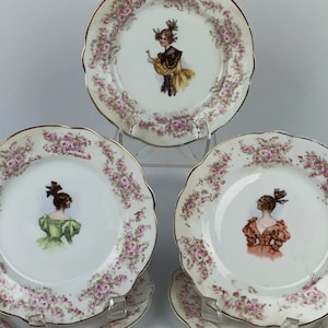 Pode incluir: Conjunto de pratos de porcelana vintage com bordas onduladas e acabamento dourado. Cada prato apresenta uma ilustração central de uma mulher em um vestido de época, cercada por uma borda de rosas cor-de-rosa. Os pratos são exibidos em suportes transparentes.