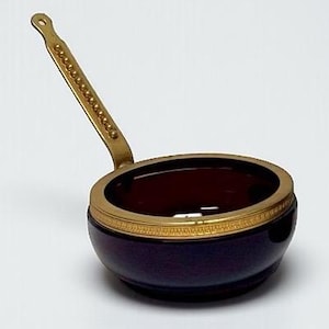 May include: A small, dark purple bowl with a gold-coloured rim and handle. The handle is long and has a series of small, decorative gold accents. The bowl is likely made of ceramic or porcelain and is designed for serving.