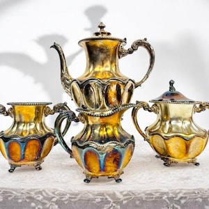 Könnte beinhalten: Ein Vintage-Teeservice mit Teekanne, Sahnekännchen und Zuckerdose. Das Set hat eine goldene Oberfläche mit aufwendigen Details und Griffen. Die Teile sind auf einer weißen Spitzentischdecke angeordnet.