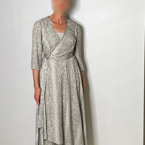 May include: A gray and white snakeskin print wrap dress with a long skirt and a high low hemline. The dress has a V-neckline and three-quarter sleeves. The model is wearing the dress with nude heeled sandals.