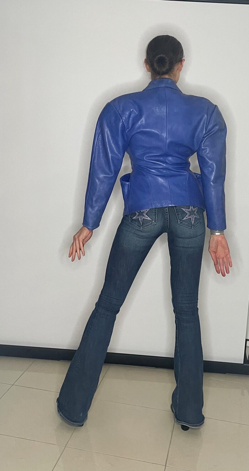 May include: A woman wearing a blue leather jacket with a cinched waist and flared jeans with star-shaped embellishments on the back pockets.