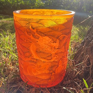 May include: A translucent orange cylindrical vessel featuring intricate relief carvings of a dragon amidst swirling clouds and waves. The surface texture is highly detailed with fine lines throughout, creating a vibrant, glowing appearance in sunlight.