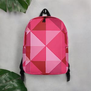 May include: A pink backpack with a geometric pattern of triangles in shades of pink and red. The backpack has black straps and a black top handle. The bag is hanging against a light gray wall.