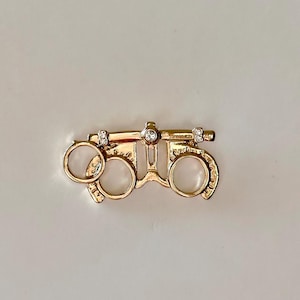May include: A gold-colored trial frame pin with clear crystal accents. The pin is designed to resemble the frame used by optometrists to test vision. The pin has two circular eye openings and a bridge with a central adjustment mechanism.