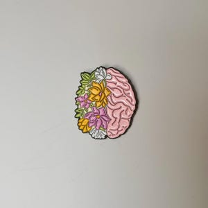 May include: A pink brain-shaped enamel pin with a floral design. The left side of the pin features colorful flowers in yellow, pink, and white, while the right side depicts the brain's structure. The pin has a black outline.