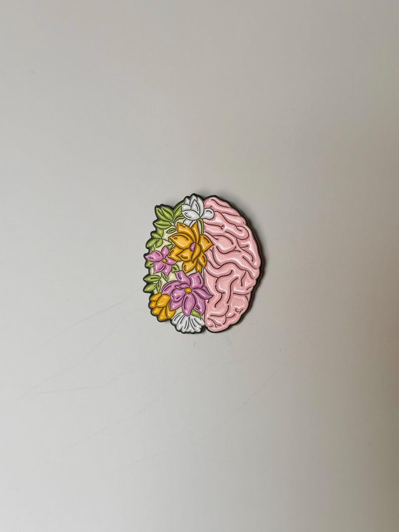 May include: A pink brain-shaped enamel pin with a floral design. The left side of the pin features colorful flowers in yellow, pink, and white, while the right side depicts the brain's structure. The pin has a black outline.