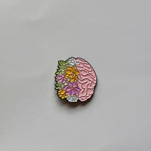 May include: A pink brain-shaped enamel pin with a floral design on the left side. The flowers are in shades of yellow, pink, white, and green. The pin has a gold outline and is set against a white background.