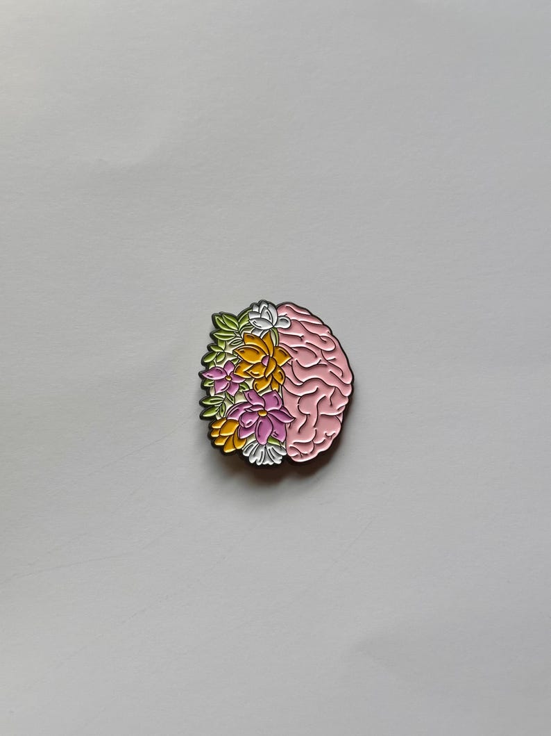 May include: A pink brain-shaped enamel pin with a floral design on the left side. The flowers are in shades of yellow, pink, white, and green. The pin has a gold outline and is set against a white background.