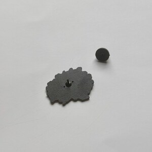 May include: A dark gray enamel pin with a floral design. The pin has a black metal backing with a pin clasp. The pin is shown on a white surface. A separate pin back is also visible.