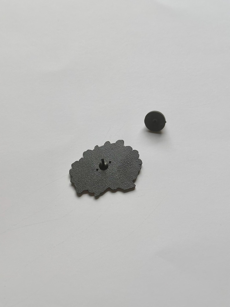 May include: A dark gray enamel pin with a floral design. The pin has a black metal backing with a pin clasp. The pin is shown on a white surface. A separate pin back is also visible.