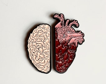 Könnte beinhalten: Zwei anatomische Emaille-Anstecknadeln, eine zeigt ein Gehirn und die andere ein Herz. Die Gehirn-Anstecknadel ist beige mit schwarzen Umrissen, und die Herz-Anstecknadel ist rot und rosa mit schwarzen Umrissen. Beide Anstecknadeln sind vor weißem Hintergrund.