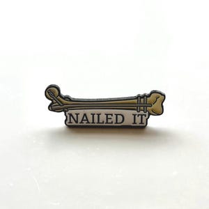 Op de afbeelding: Een zwart-witte emaille pin met een cartoon afbeelding van een gebroken bot met een spijker erdoorheen en de tekst "NAILED IT".