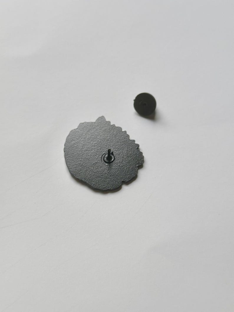 May include: A dark gray enamel pin with a floral design, and a separate pin back. The pin has a black metal clasp on the back. The pin is on a white surface.