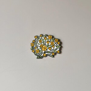 May include: A brain-shaped brooch with a white base, adorned with yellow flowers and green leaves. The brooch has a silver-colored outline. The design is intricate and detailed, suggesting a nature or botanical theme.