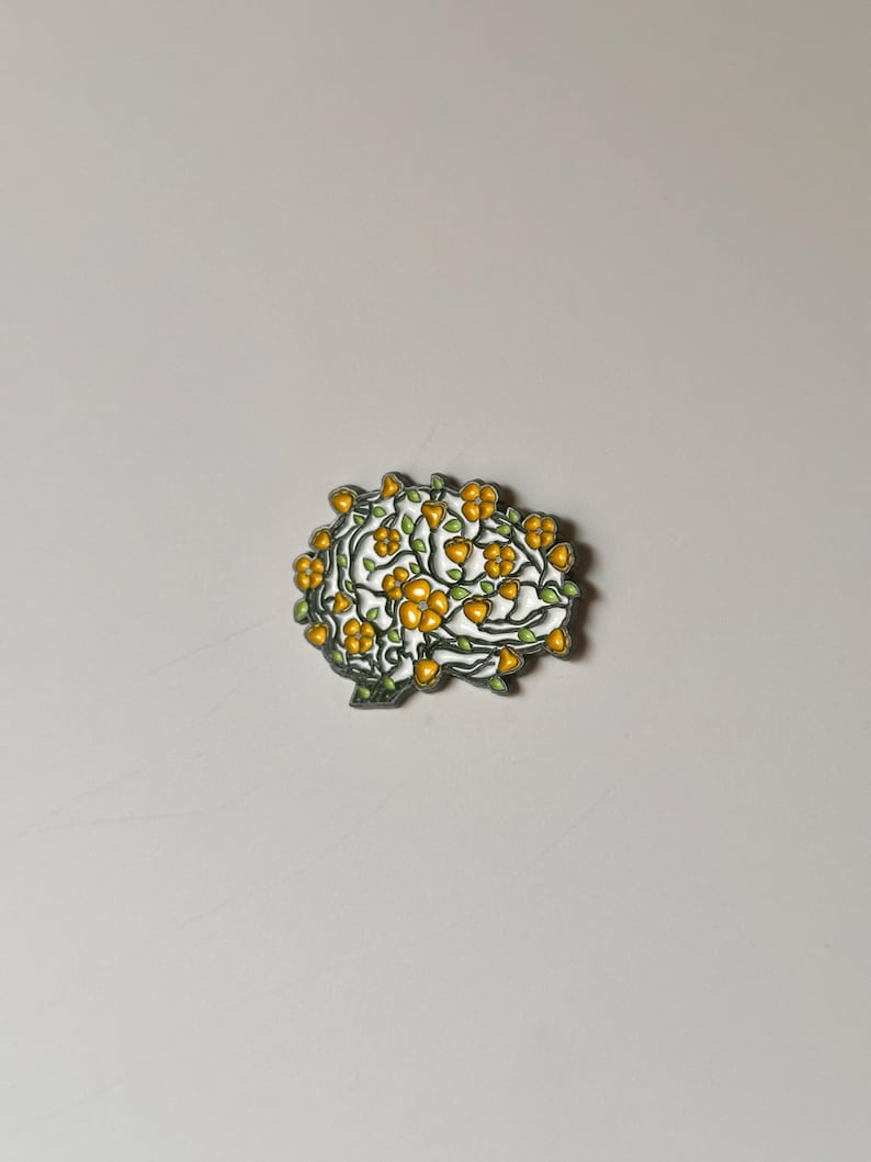 May include: A brain-shaped brooch with a white base, adorned with yellow flowers and green leaves. The brooch has a silver-colored outline. The design is intricate and detailed, suggesting a nature or botanical theme.