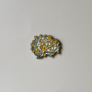 May include: A brain-shaped enamel pin with a floral design. The pin features a white outline with yellow flowers and green leaves. The pin is set against a plain, light gray background.
