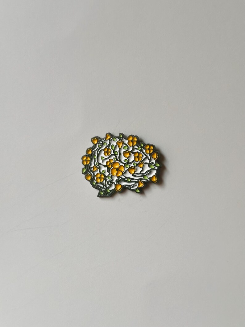May include: A brain-shaped enamel pin with a floral design. The pin features a white outline with yellow flowers and green leaves. The pin is set against a plain, light gray background.