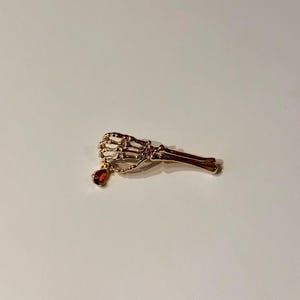 May include: A gold-coloured skeleton hand brooch with a dangling red gemstone. The brooch features articulated fingers and a delicate chain. The design is a unique, gothic-inspired accessory, suitable for adding a touch of the macabre.