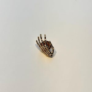 Könnte beinhalten: Eine goldfarbene Anstecknadel in Form einer Skeletthand mit detaillierten Fingerknochen. Die Nadel ist vor einem weißen Hintergrund platziert, der ihr aufwendiges Design hervorhebt. Dieses Accessoire eignet sich, um Kleidung oder Accessoires eine einzigartige Note zu verleihen.