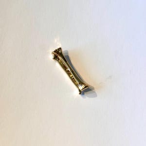 May include: A gold-colored, elongated bar-shaped jewelry piece with a decorative design. The piece has a slight curve and is set against a white background. The jewelry appears to be made of metal.