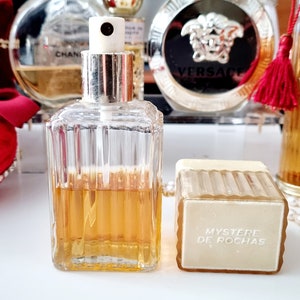May include: A clear glass bottle of perfume with a gold cap and a white spray nozzle. The bottle is filled with a light amber liquid. The bottle is sitting on a white surface next to a white and gold box with the text "Mystere de Rochas" printed on it.
