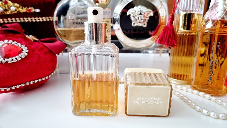 May include: A clear glass bottle of perfume with a gold cap and a white spray nozzle. The bottle is filled with a light amber liquid. The bottle is sitting on a white surface next to a white and gold box with the text "Mystere de Rochas" printed on it.
