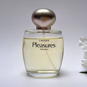 Może przedstawiać: Przezroczysta szklana butelka perfum z srebrną zakrętką. Butelka jest oznaczona etykietą "Lauder Pleasures for Men".