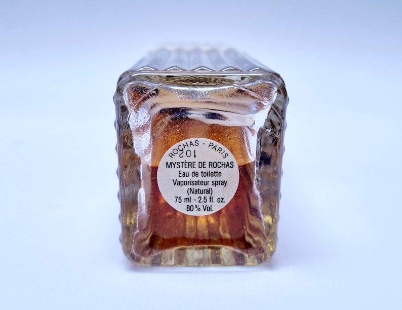 May include: A clear glass bottle of perfume with a brown label that reads "ROCHAS PARIS MYST&Egrave;RE DE ROCHAS Eau de toilette Vaporisateur spray (Natural) 75 ml - 2.5 fl. oz. 80% Vol."