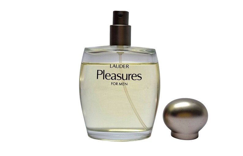 Może przedstawiać: Przezroczysty szklany flakon perfum z złotą zakrętką i srebrnym atomizerem. Etykieta zawiera napis "LAUDER Pleasures FOR MEN".