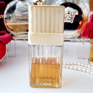 May include: A clear glass bottle of perfume with a gold cap and a white label. The bottle is filled with a light amber liquid. The bottle is sitting on a white surface.